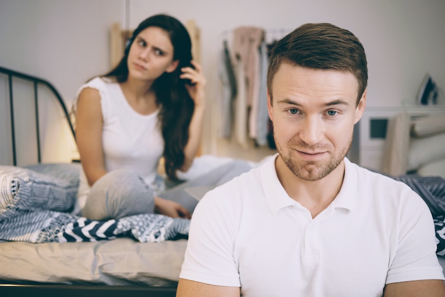 Unglückliche Frau sitzt auf einem Ehebett, während ein Mann mittleren Alters im Raum ist – Darstellung eines vulnerablen Narzissten und seiner Auswirkung auf die Sexualität