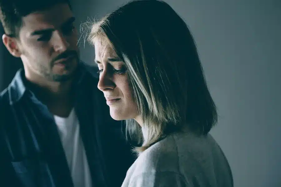 Eine junge Frau schaut traurig zu Boden, während ein Mann im Hintergrund besorgt zu ihr blickt – eine emotionale Szene, die Schmerz und Distanz in einer Beziehung zeigt