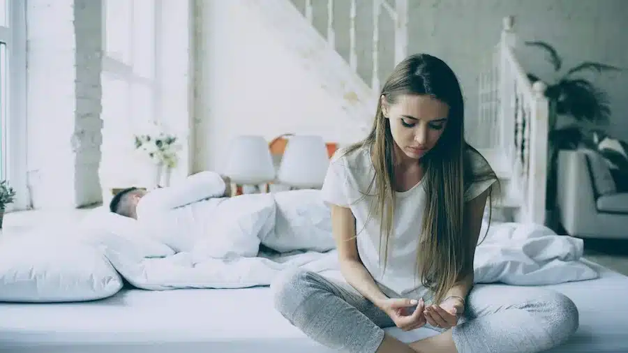 Eine junge Frau sitzt nachdenklich am Rand eines Bettes und schaut auf ihre Hände, während ein Mann im Hintergrund im Bett liegt – eine Szene, die emotionale Distanz und Traurigkeit in einer Beziehung ausdrückt