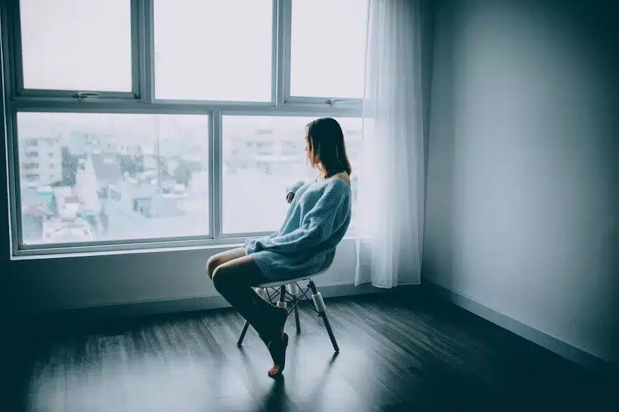 Frau sitzt mit dem Rücken zur Kamera auf einem Baumstamm am Meer und blickt ruhig auf das Wasser und den Horizont