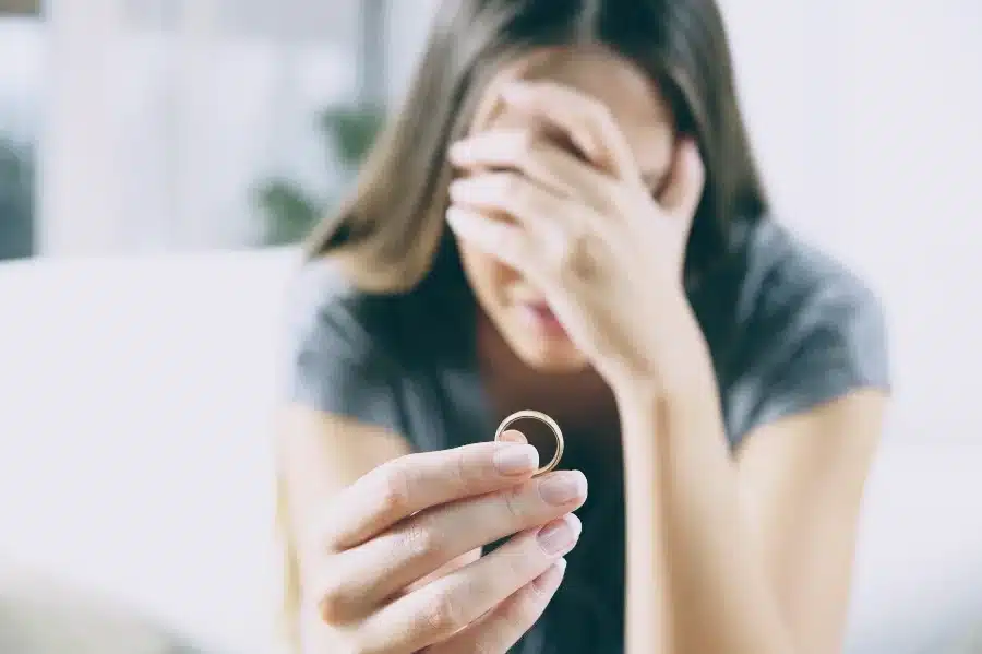 Traurige Frau sitzt nachdenklich auf dem Sofa und hält ihren Ehering in der Hand – Symbolbild für Trennung, Scheidung oder Loslassen