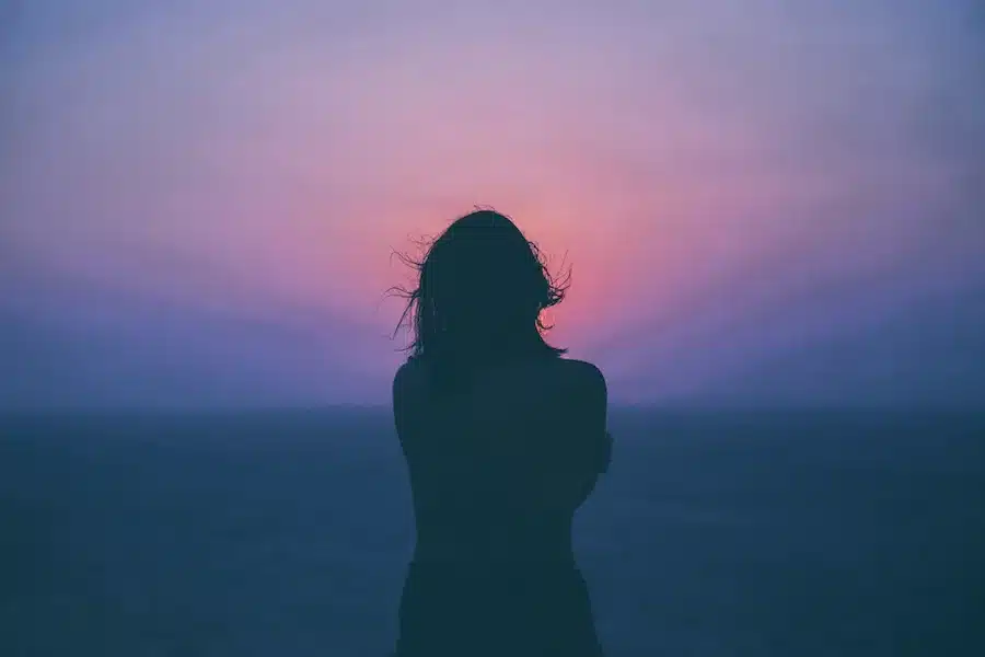 Silhouette einer Frau, die allein am Meer steht, vor einem sanften Sonnenuntergang in Rosa- und Blautönen, mit leicht im Wind wehenden Haaren, in einer ruhigen, nachdenklichen Stimmung