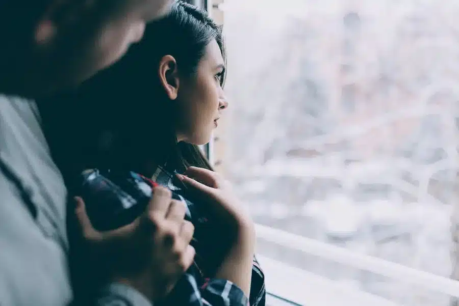 Frau blickt nachdenklich aus dem Fenster, während ein Mann hinter ihr Nähe sucht – Darstellung emotionaler Distanz und Nichtverfügbarkeit in Beziehungen