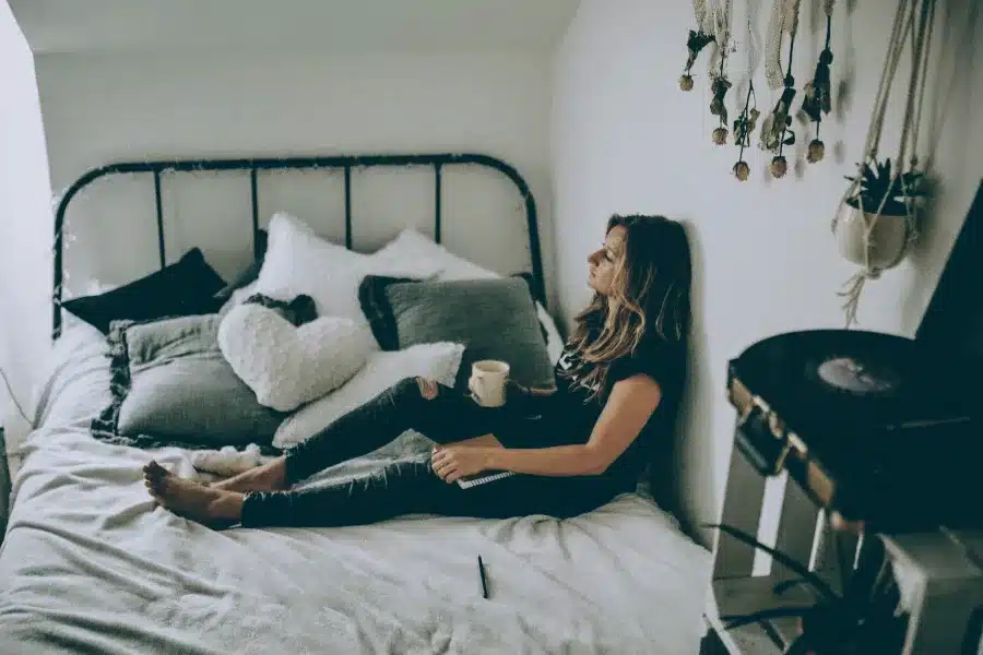 Eine junge Frau sitzt nachdenklich auf einem Bett, umgeben von Kissen, hält eine Tasse in der Hand und schaut an die Wand. Das Zimmer wirkt gemütlich und ruhig, mit hängenden Pflanzen und einem Plattenspieler im Hintergrund