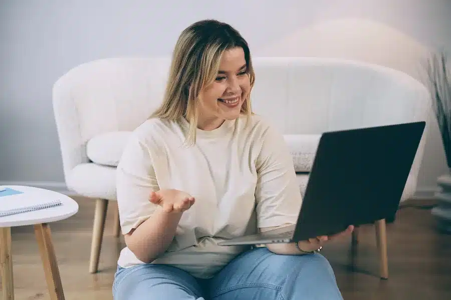 Junge Frau in Jeans hält einen Laptop in der Hand und führt einen Videoanruf, lächelnd und konzentriert, vor einem hellen Hintergrund