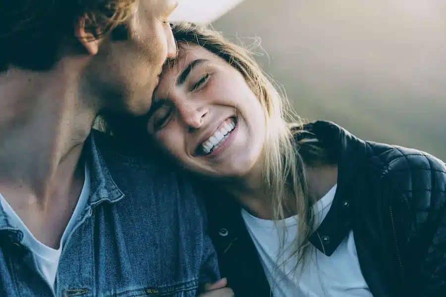 Ein junges Paar sitzt nah beieinander. Der Mann küsst sanft die Stirn der Frau, die mit geschlossenen Augen und einem breiten Lächeln an seine Schulter gelehnt ist. Beide wirken glücklich und vertraut, draußen in natürlichem Licht. Sie trägt eine schwarze Lederjacke, er eine blaue Jeansjacke über einem weißen T-Shirt